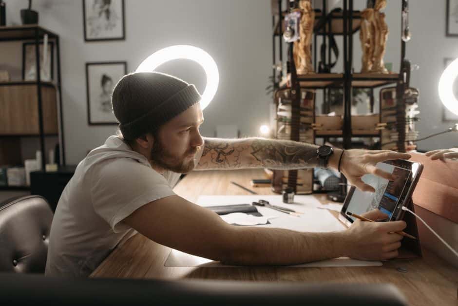 A tattooed man wearing a beanie sketching on a tablet at a wooden table in an art studio.