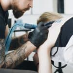 Tattoo artist with beard and gloves creating a back tattoo in a studio setting.