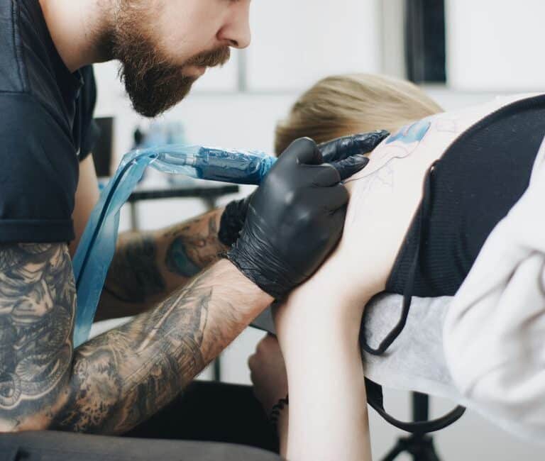 Tattoo artist with beard and gloves creating a back tattoo in a studio setting.